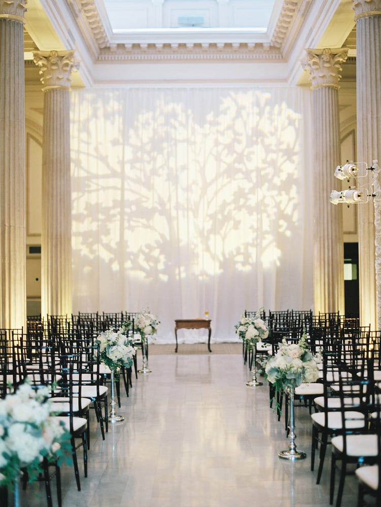 A long, elegant white drape is the backdrop for this classic wedding ceremony decor with tall floral arrangements lining the aisle.