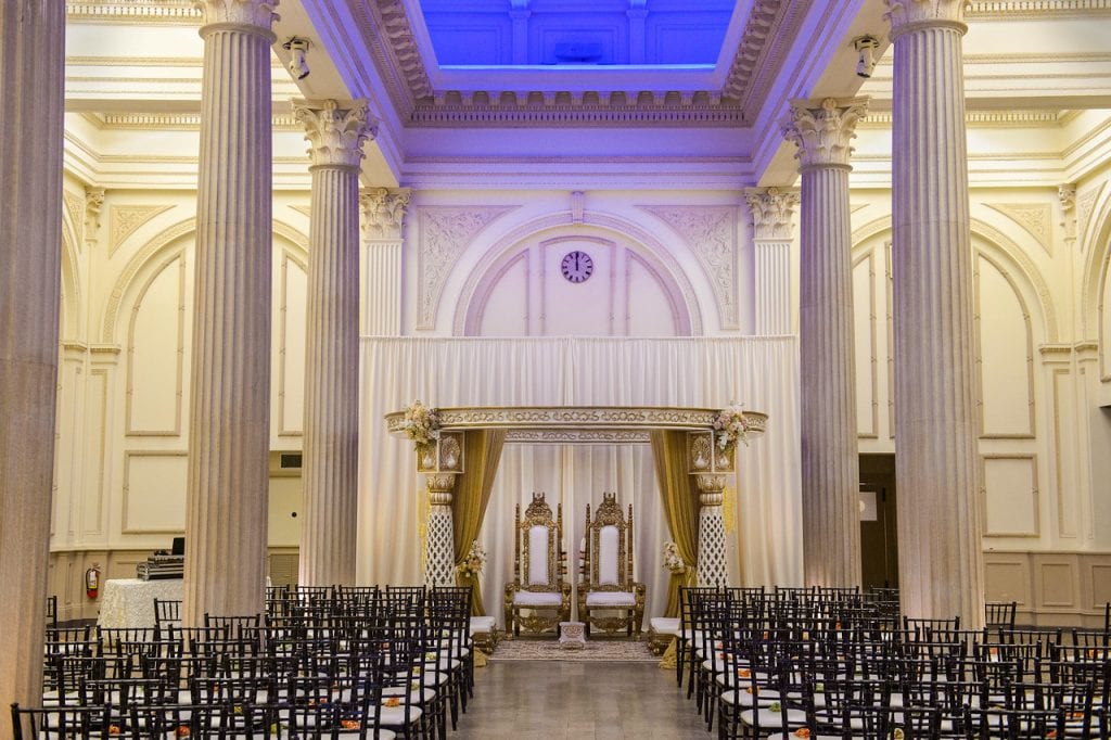 Two gold thrones with white upholstery and a white curtain backdrop mark the altar for the traditional Indian wedding ceremony.