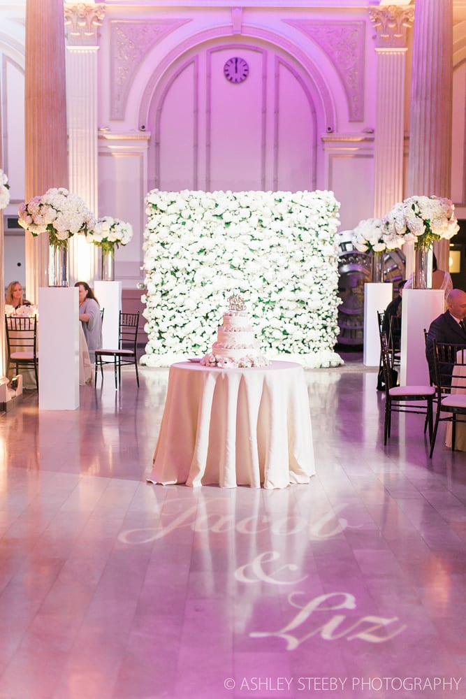 Tables and chairs are moved to the side of the room for the reception with the flower wall in the background and elegant wedding cake taking center stage.