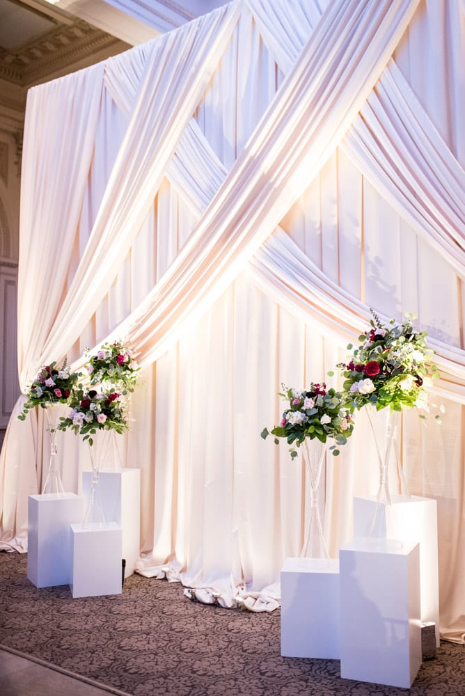 Tall floral arrangements sit on white blocks in front of white drapery for an elegant wedding ceremony backdrop.