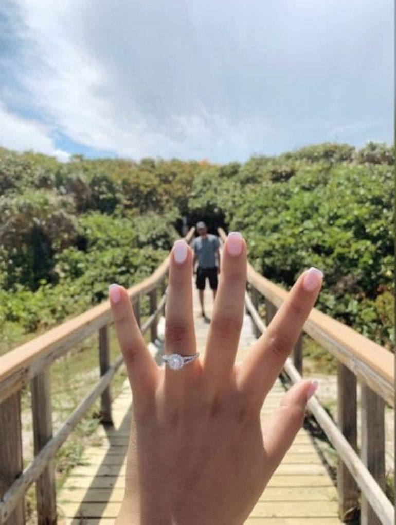 A woman shows off her new engagement ring on a bridge with her fiancé in the background.