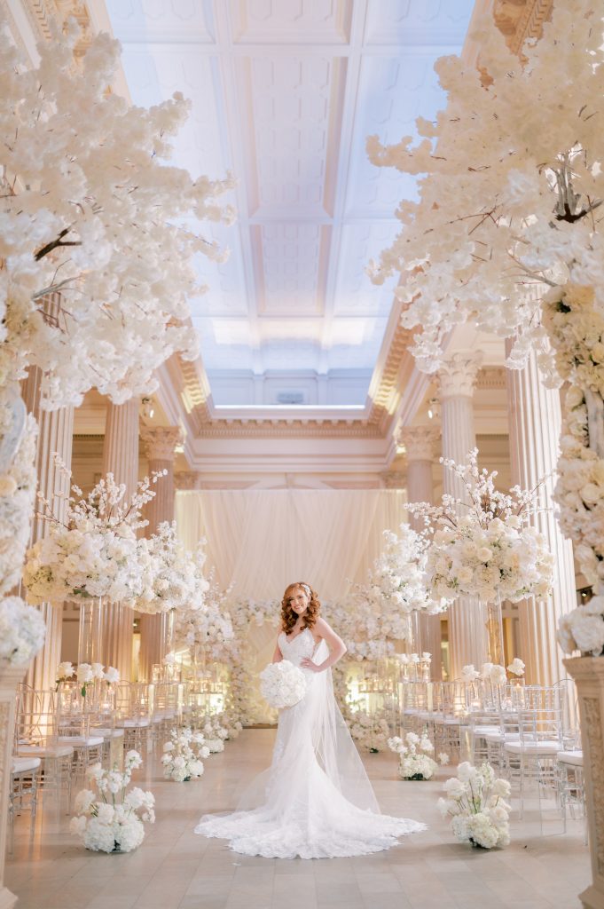 Bride in wedding aisle decorated in all white flowers with towering cherry blossom branches